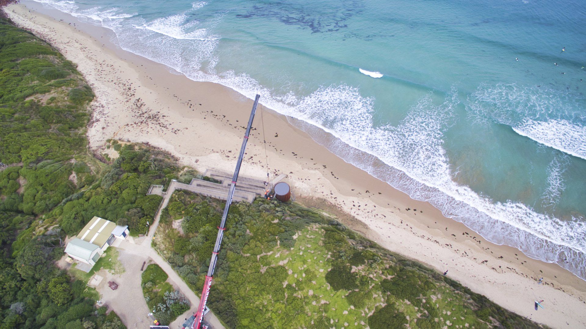 Aerial view of a beach with waves, green vegetation, and a long pier extending into the water.
