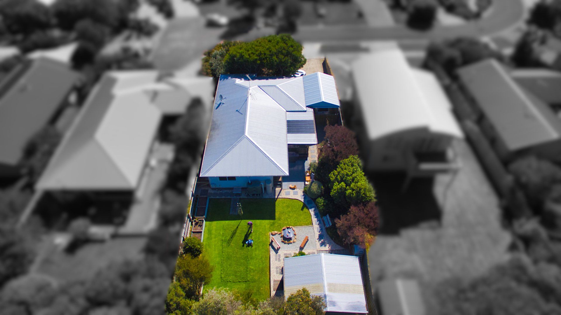 Aerial view of a suburban house with a bright blue roof and green backyard, surrounded by blurred neighbors