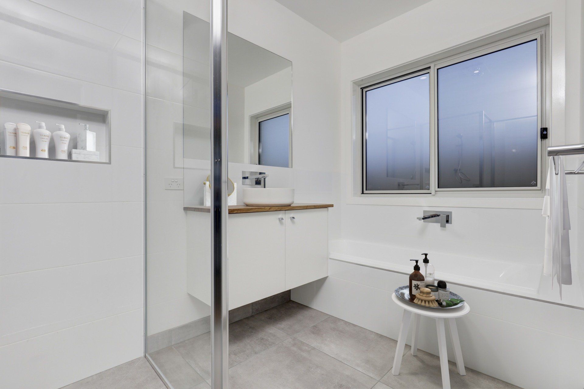 Bright white bathroom with glass shower, vanity, window, and small stool by the tub.