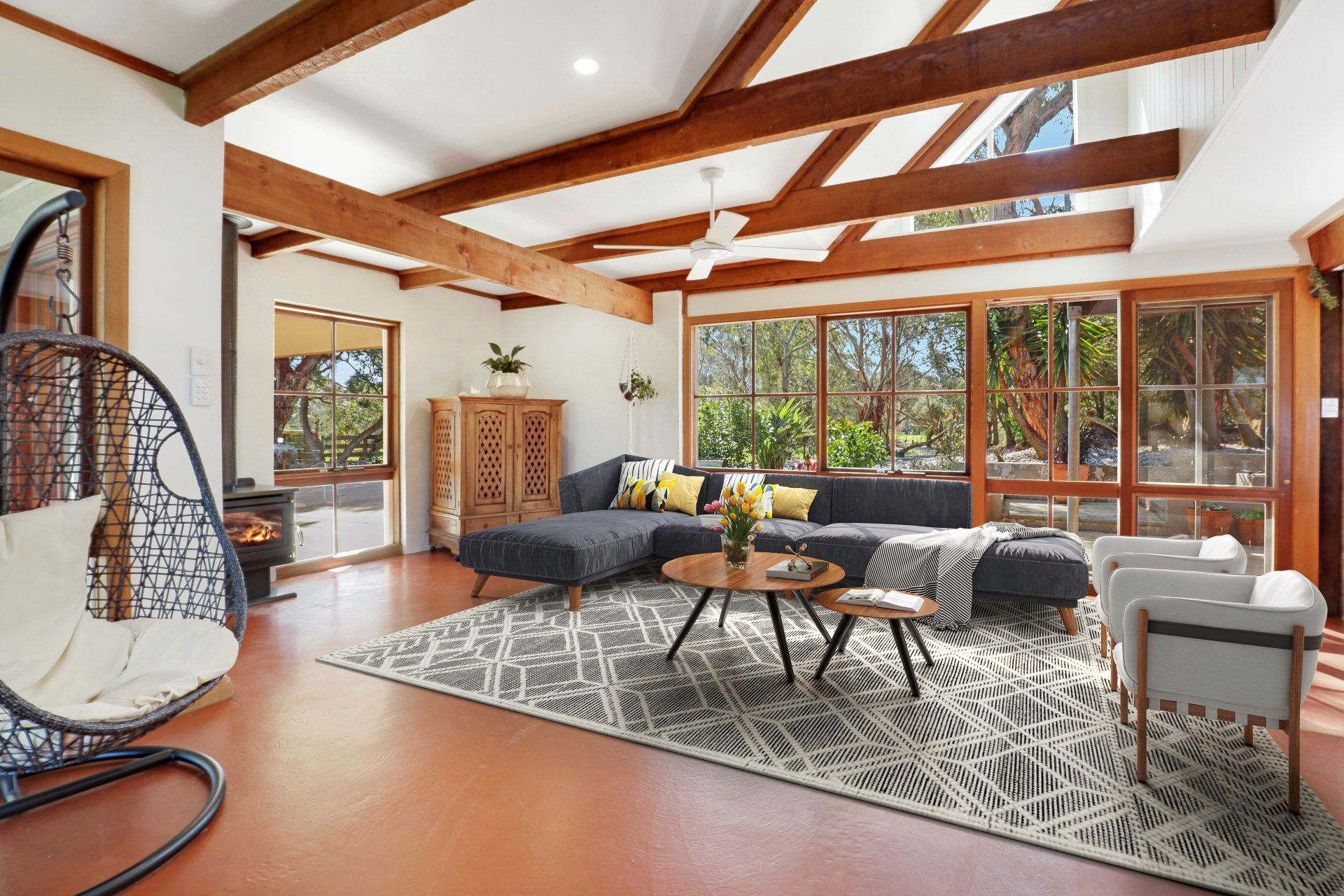 Modern living room with exposed wood beams, large windows, gray sectional, patterned rug, and hanging chair