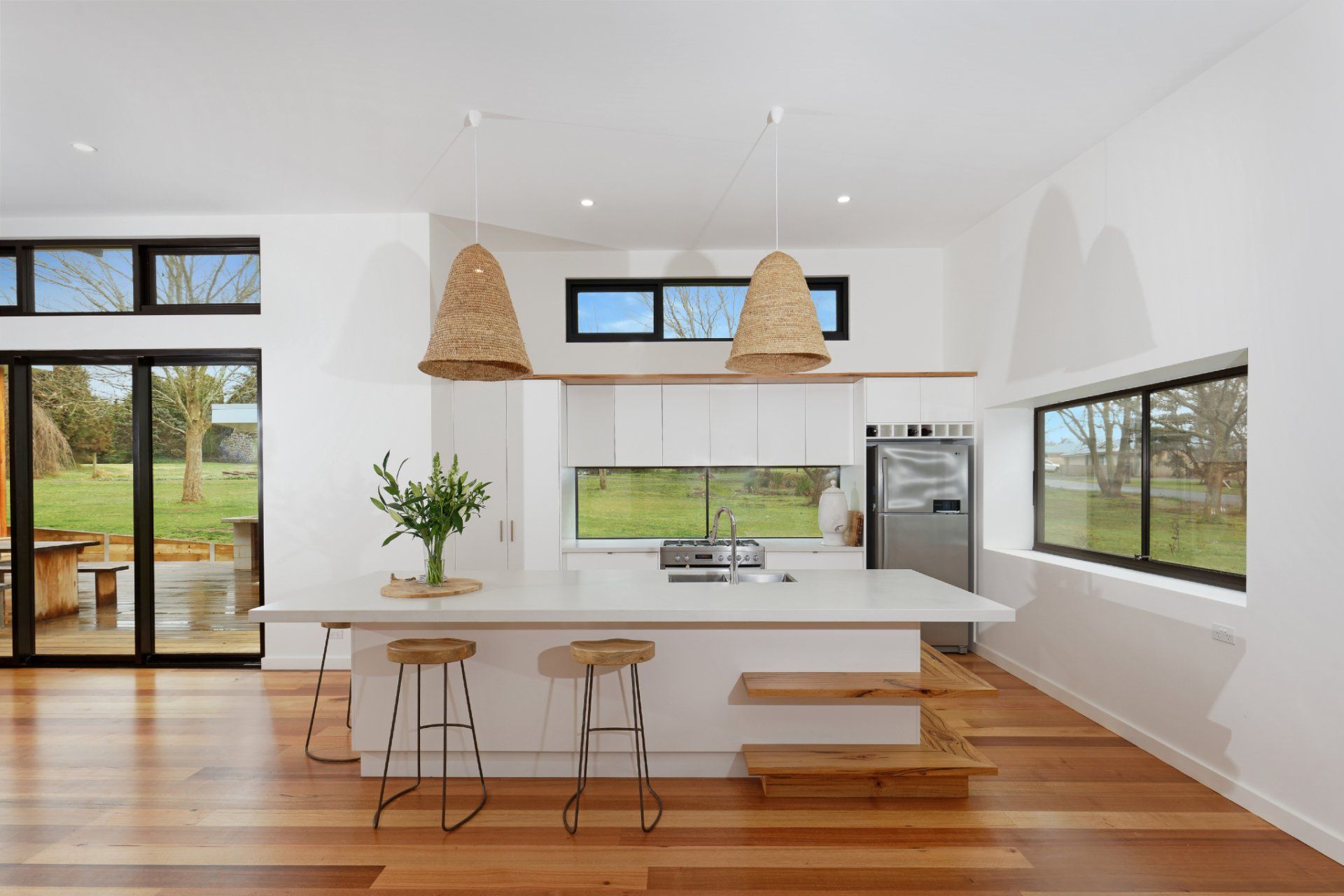 Bright modern kitchen with white cabinets, wood floors, island stools, and large windows overlooking greenery