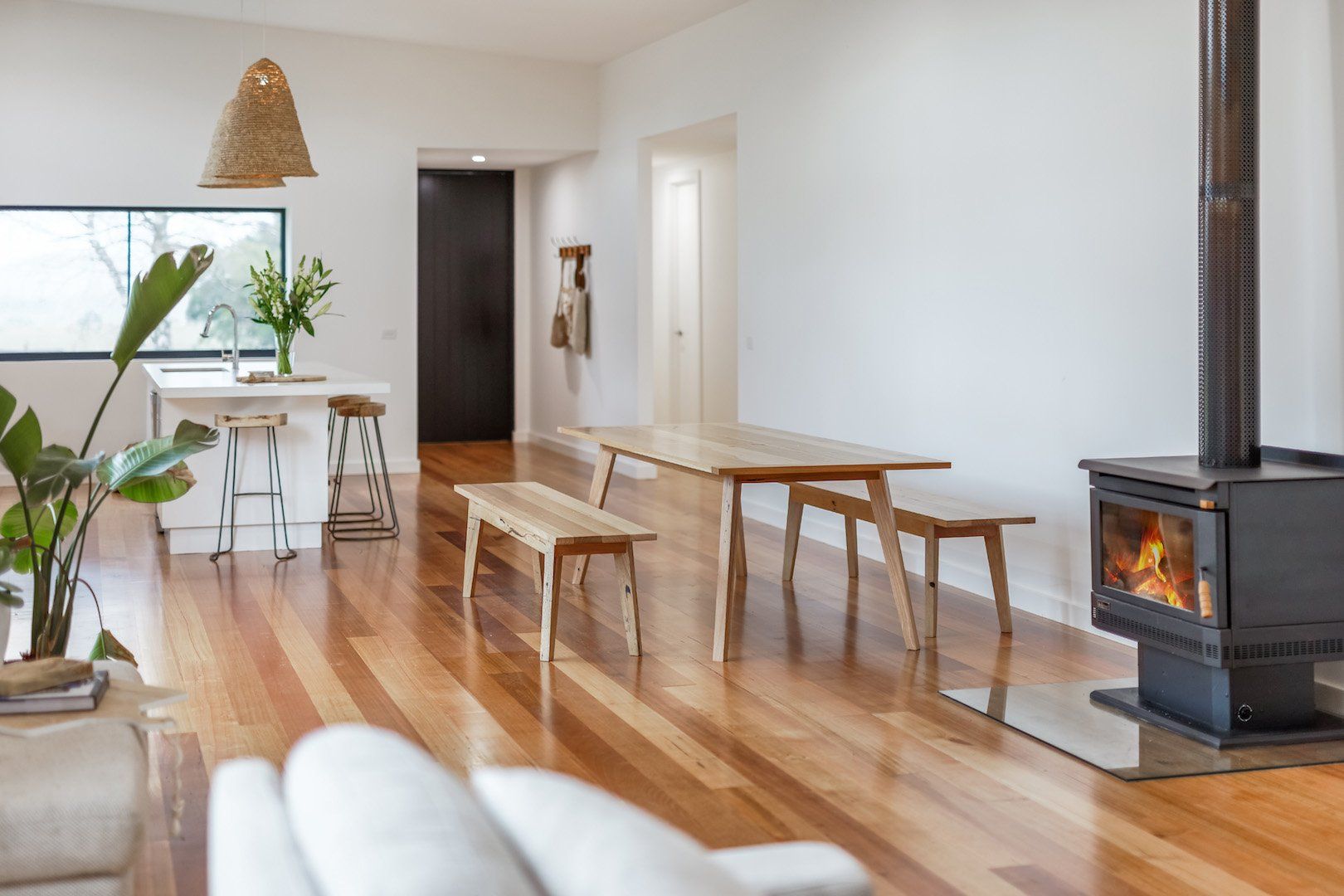 Modern open-plan living room with wood floor, long bench table, and a black wood stove by a window.