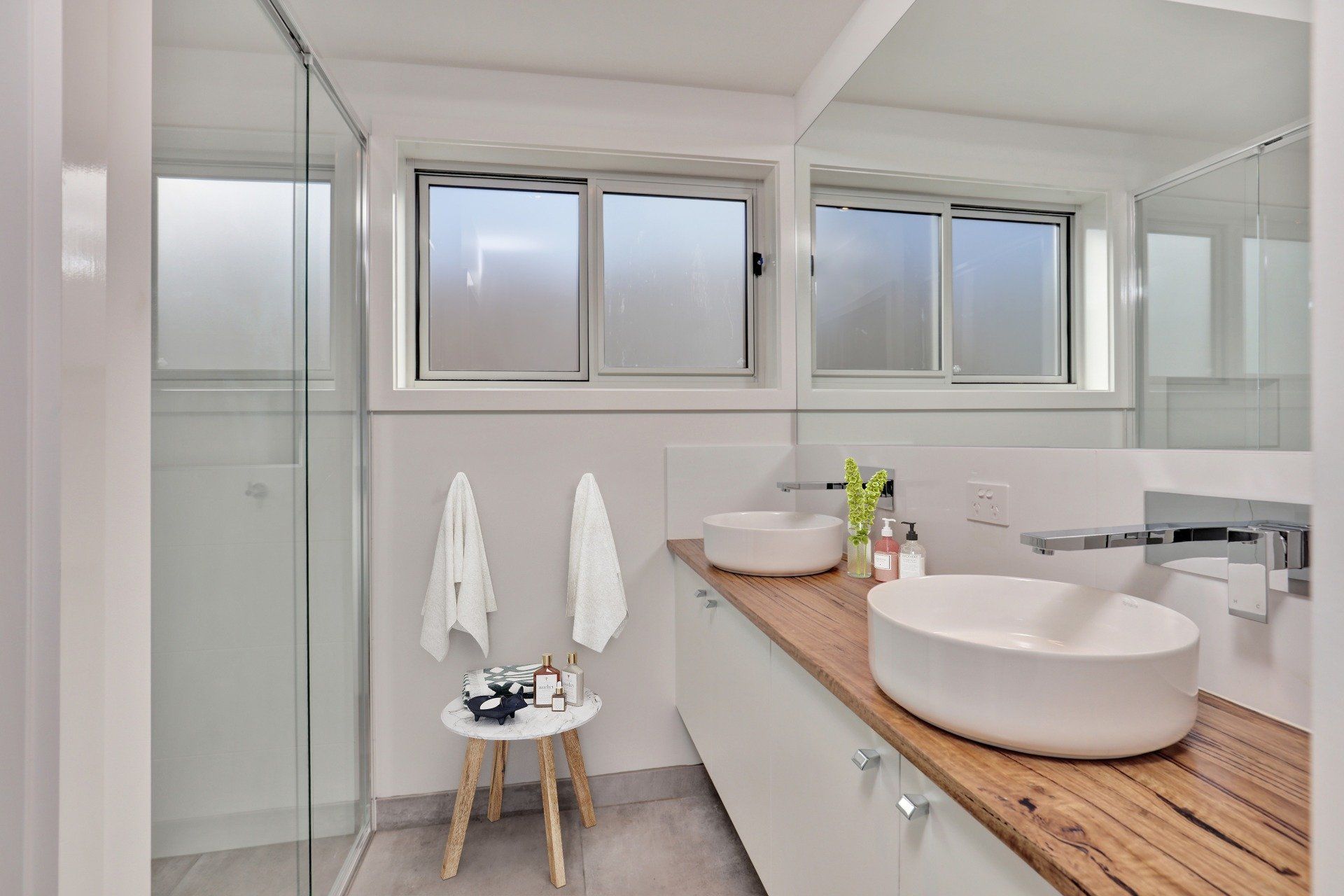 Bright modern bathroom with glass shower, double sink vanity, and towels on a stool
