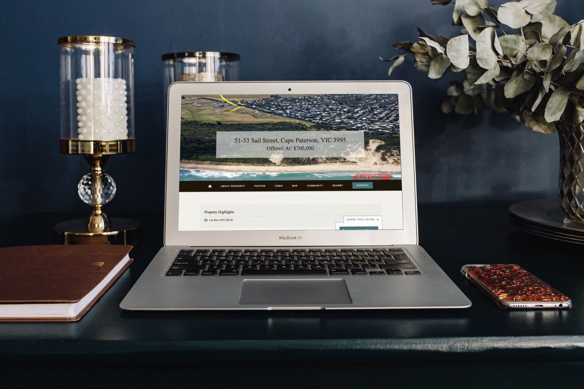 Laptop on a dark desk with a website open, flanked by a lamp, notebook, flowers, and a phone.