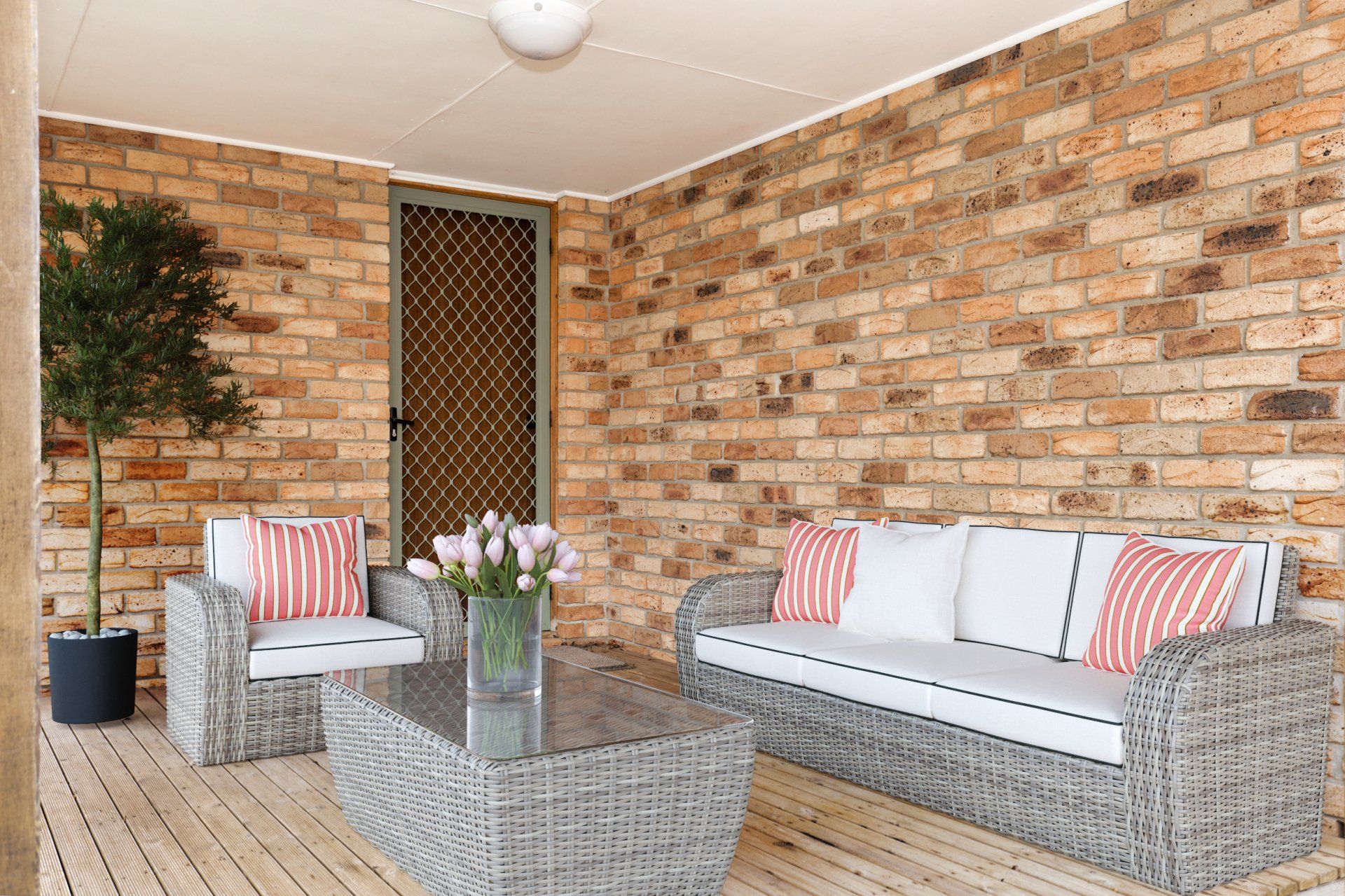 Covered patio seating area with wicker sofa, coffee table, potted tree, and brick wall backdrop
