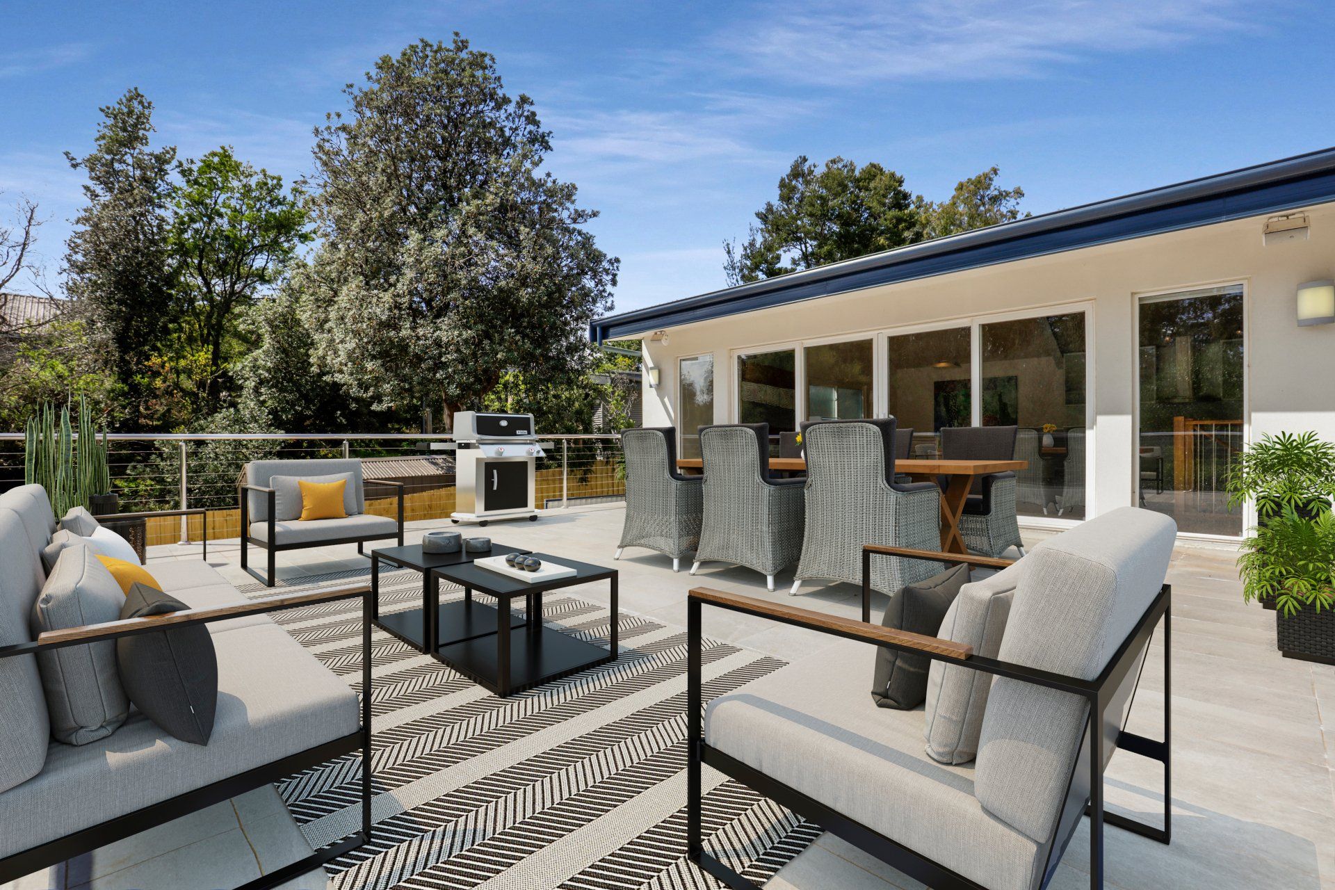 Modern patio with gray seating, patterned rug, and outdoor kitchen beside a house under a blue sky