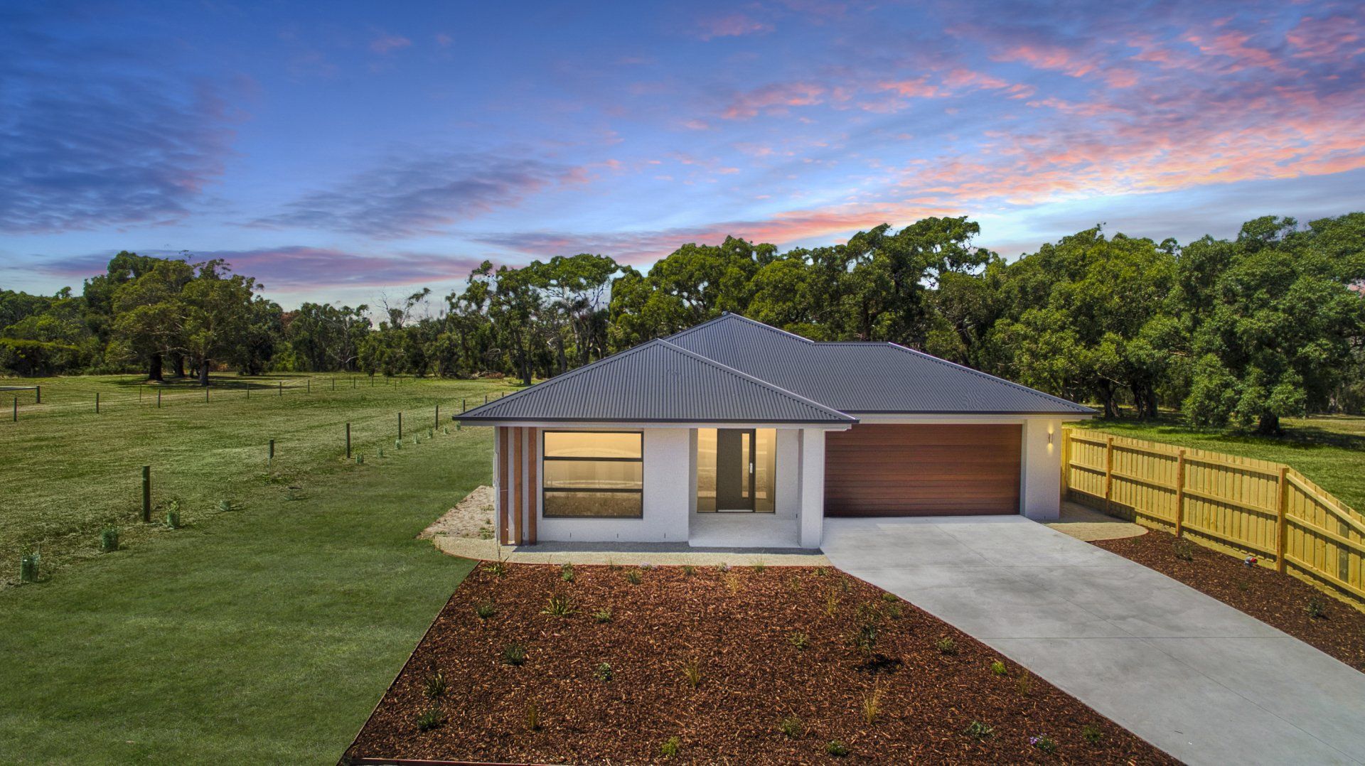 Modern single-story house with a gray roof, lit windows, a driveway, and grassy yard at dusk