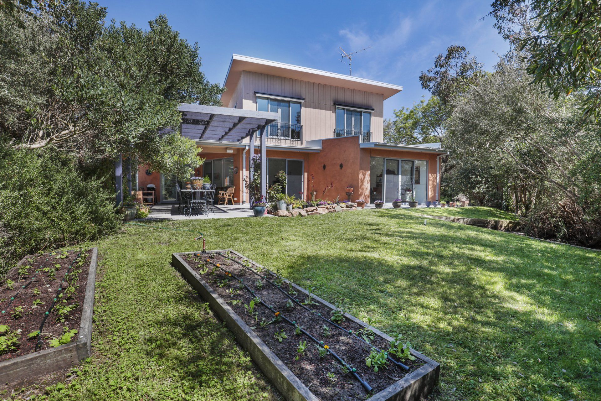 Modern two-story house with a grassy yard, raised garden beds, and trees under a blue sky