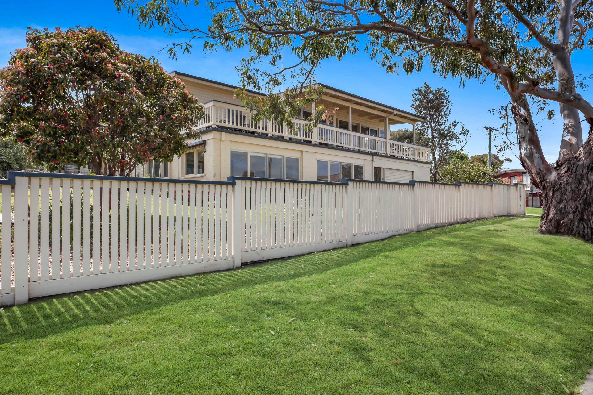 White picket fence in front of a beige two-story house with a green lawn and trees under a blue sky