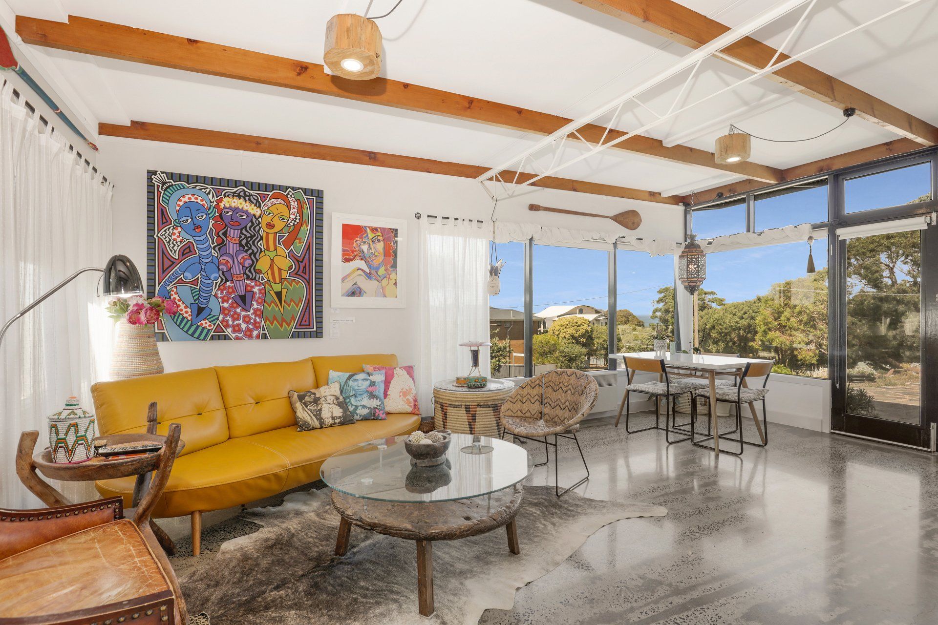 Bright living room with yellow sofa, colorful art, dining table, and large windows overlooking the hills