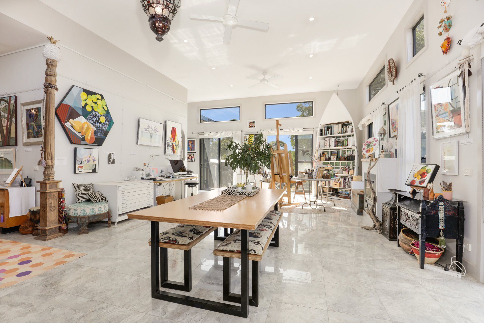 Bright art-filled studio with a long wooden table, white walls, and large windows opening to a sunny patio.