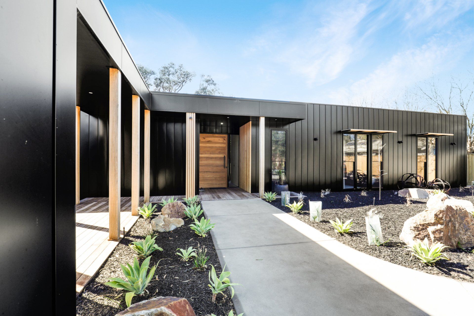 Modern black house with a concrete walkway, landscaped front garden, and wooden front door