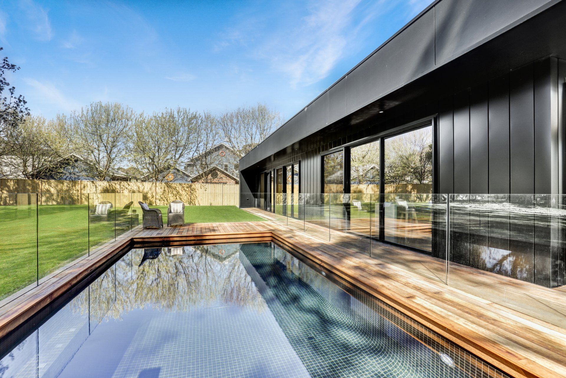 Modern house with a long reflecting pool, wood deck, and large glass walls beside a grassy yard
