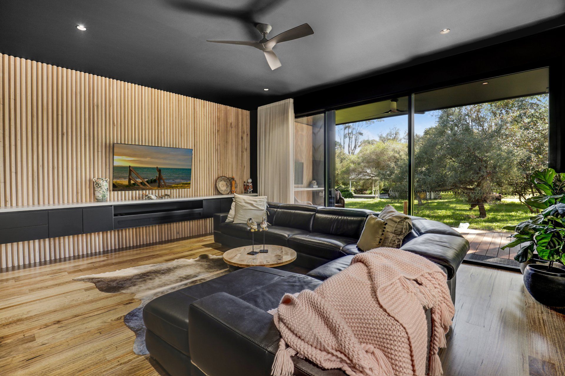 Modern living room with black sectional, wood accent wall, and large windows overlooking greenery