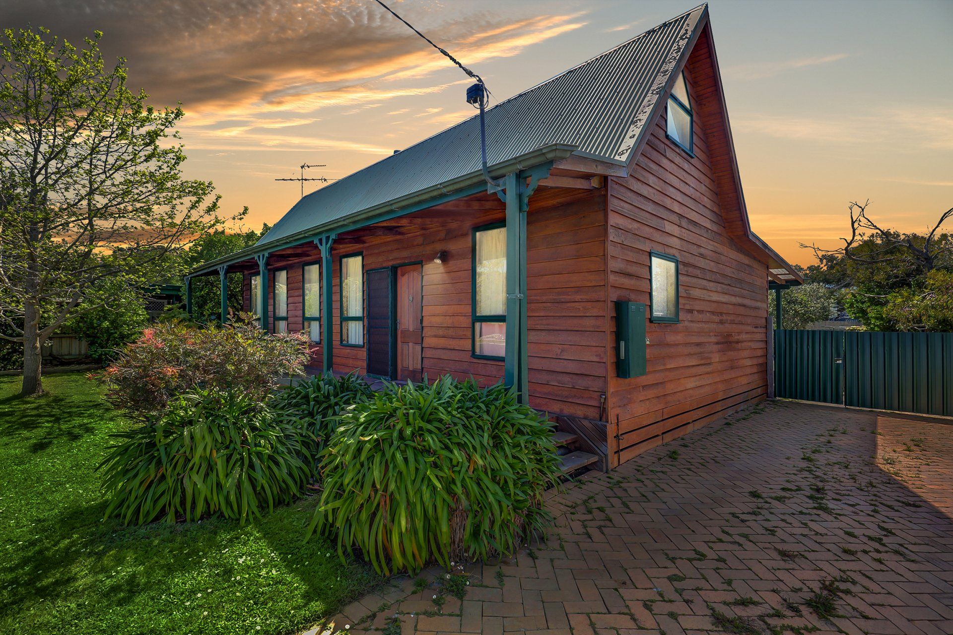 Brick house with a sloped metal roof at sunset, surrounded by greenery and a paved driveway.