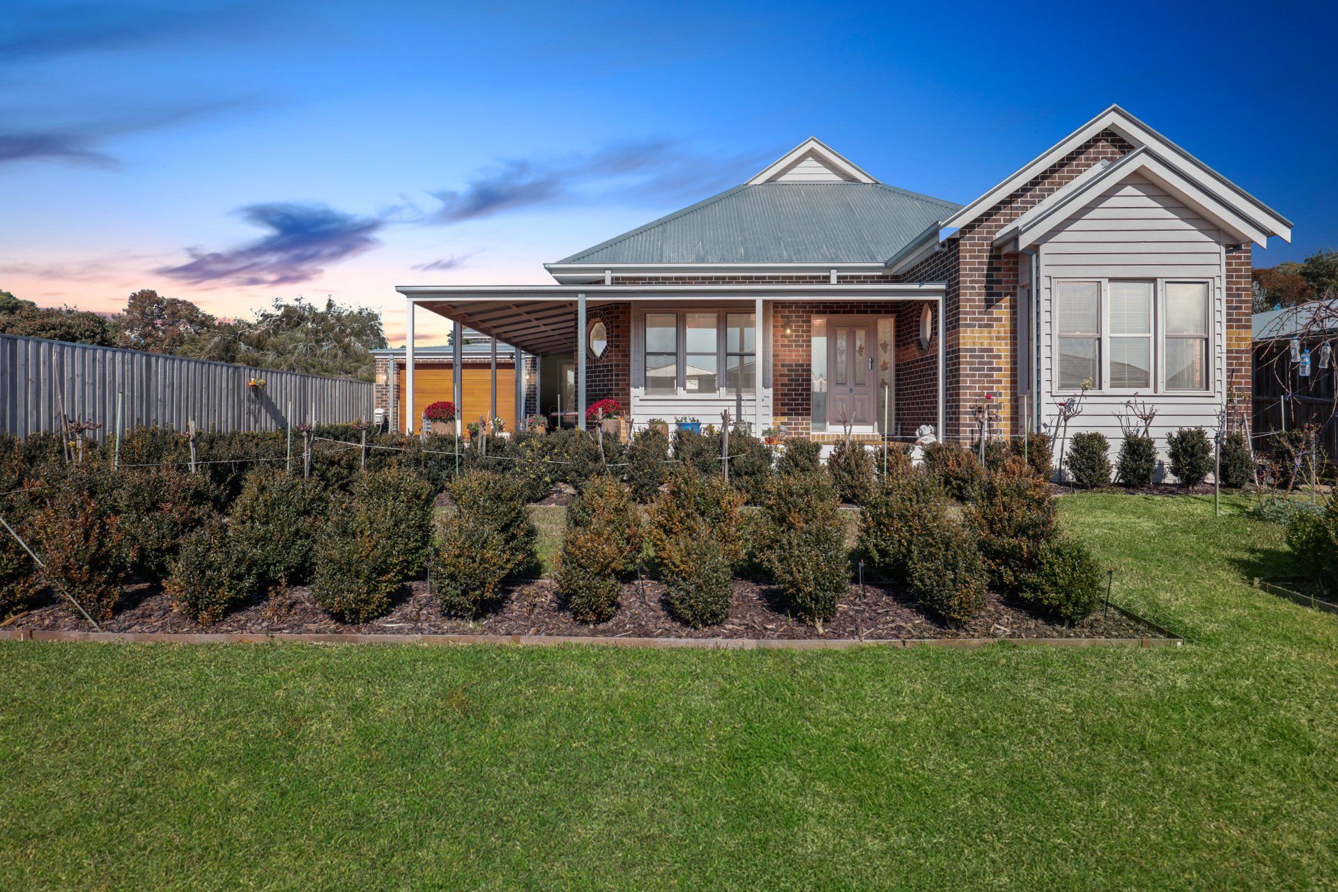 Suburban brick house with a covered porch, manicured shrubs, and a green lawn at sunset
