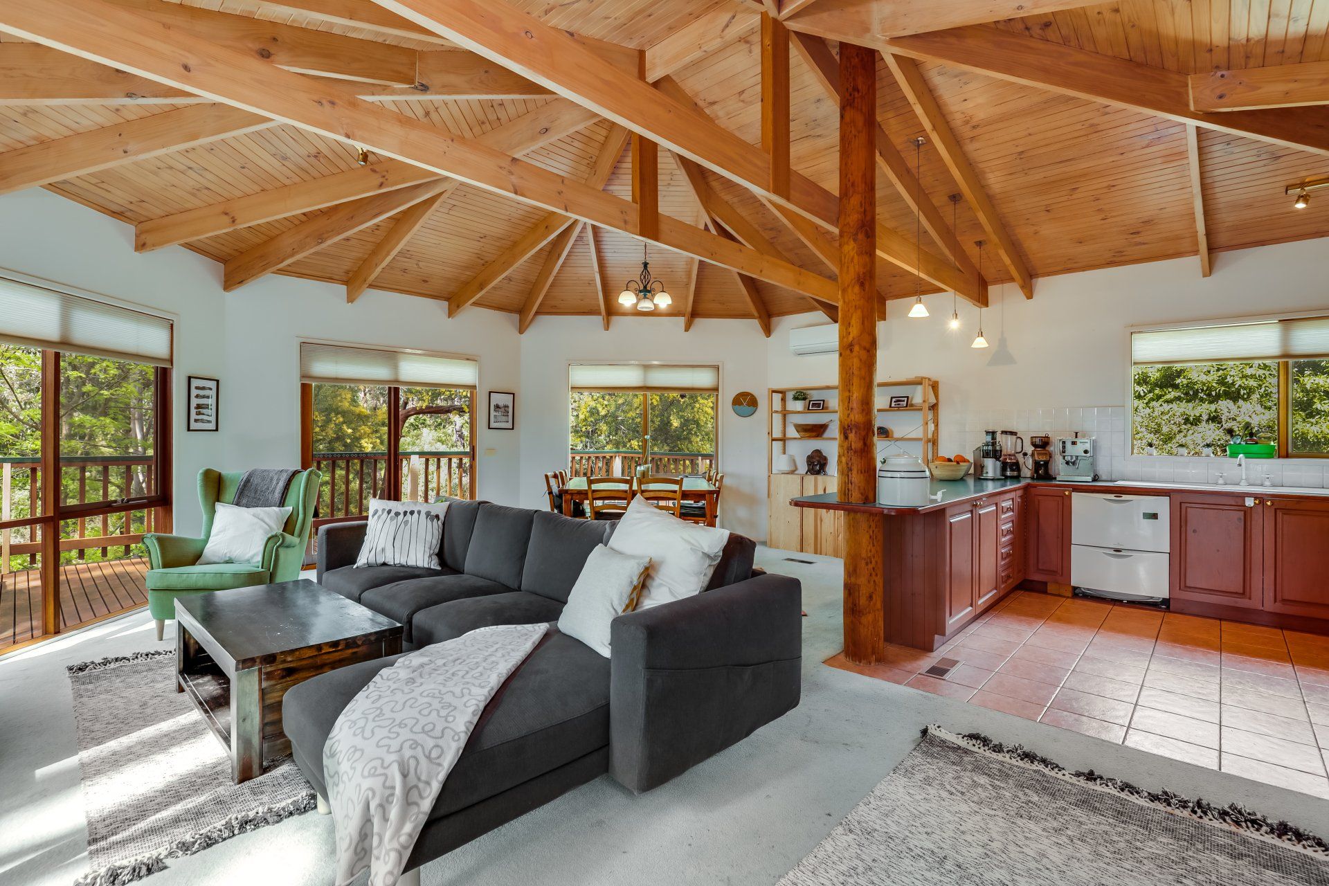 Bright open living room with wood-beamed ceiling, dark sofa, and kitchen area by large windows