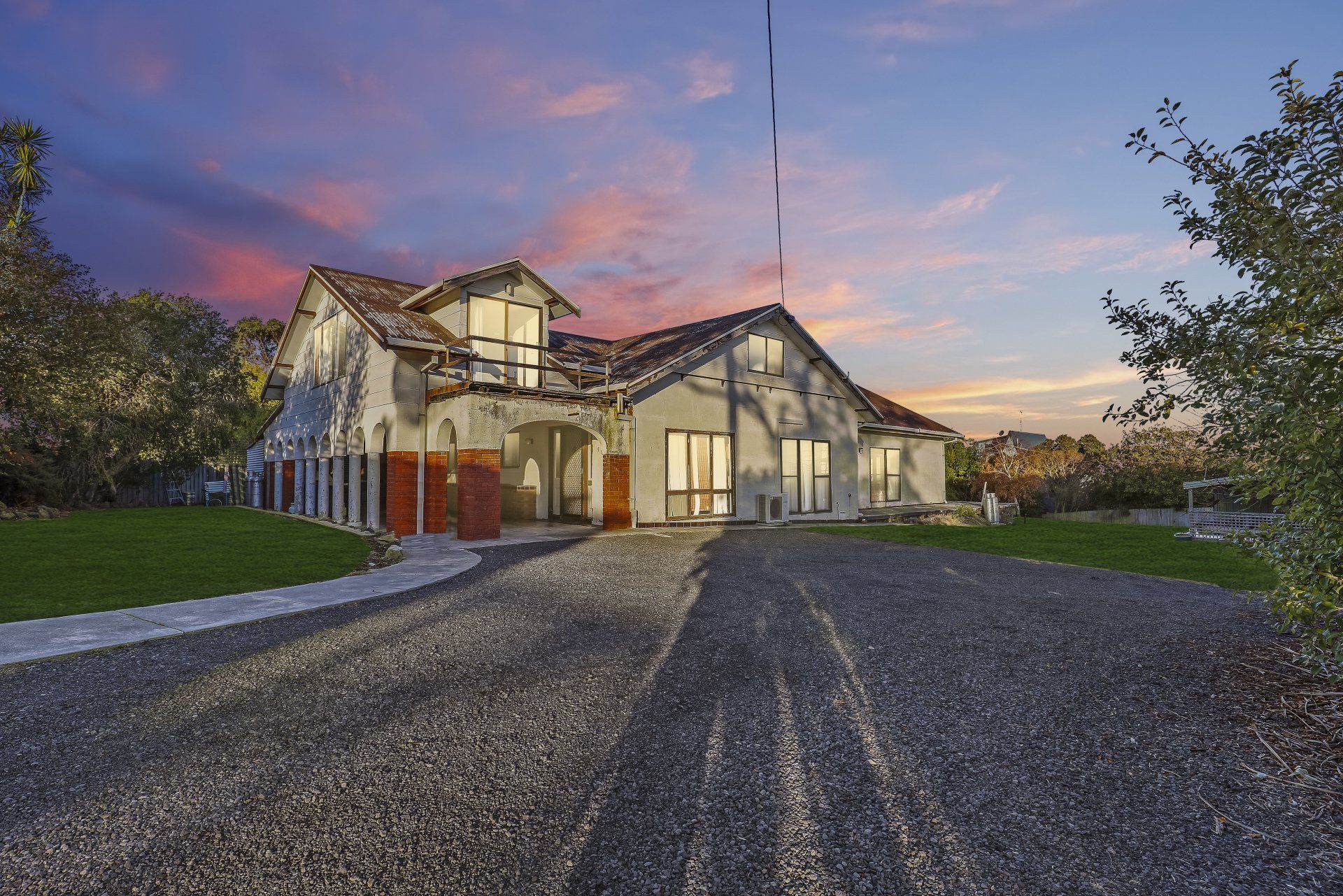 Large stone house with warm lights at dusk, seen from a gravel driveway under a pink-blue sky