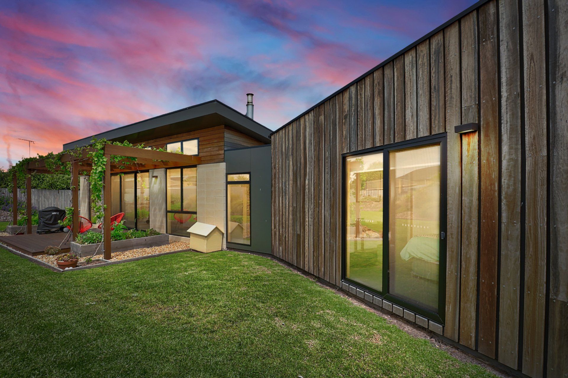 Modern house exterior at dusk with wood siding, glass doors, and a green lawn under a colorful sky