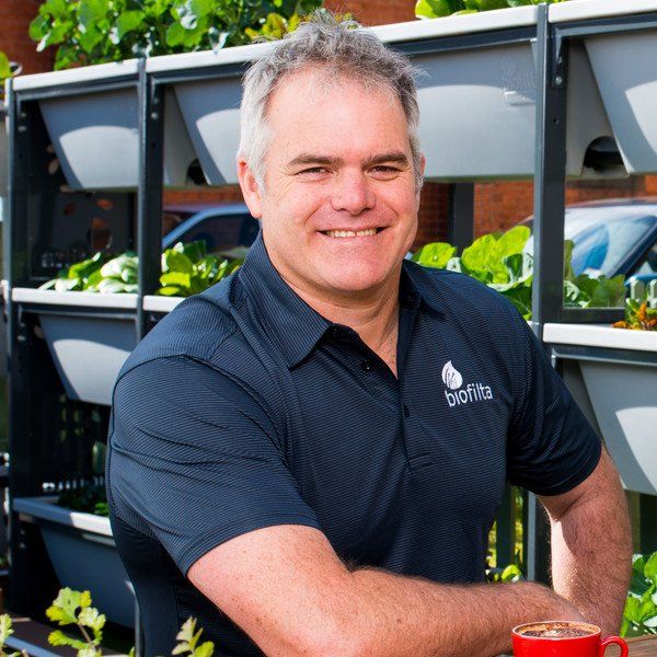 Smiling man in a dark polo stands beside a vertical herb garden outdoors