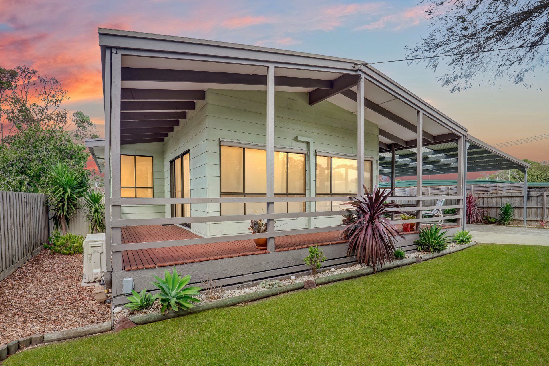 Modern single-story house with a covered porch, white railing, and a green lawn at sunset