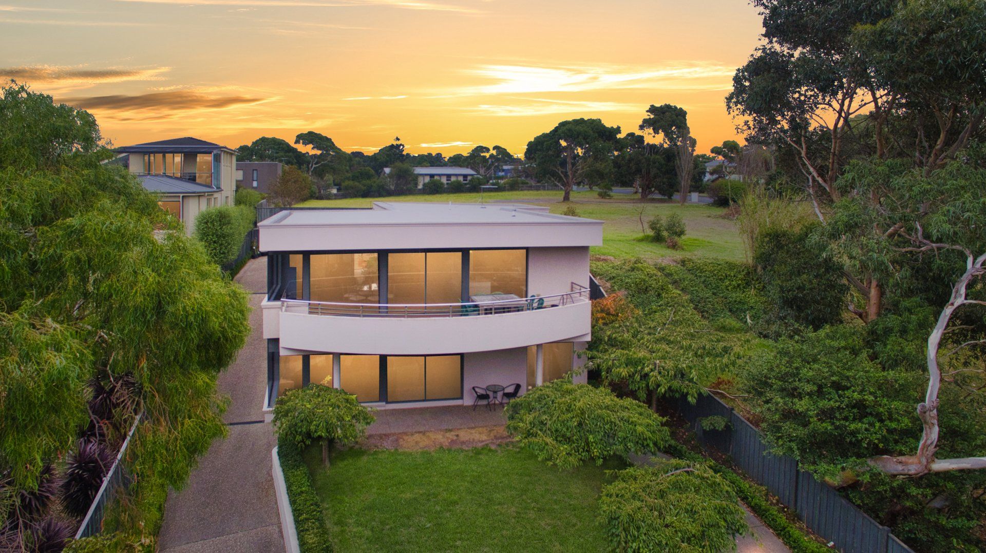 Modern two-story house with a curved facade at sunset, surrounded by greenery and a driveway