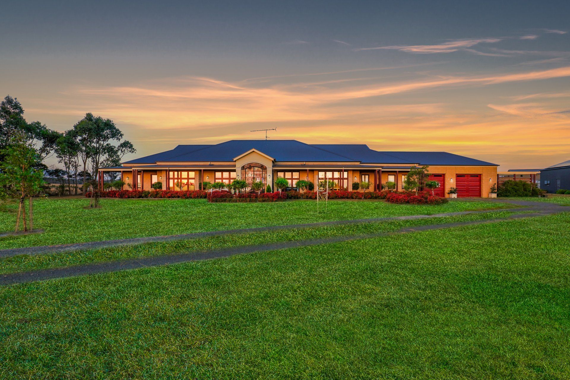 A red-brick building at sunset with lit windows, a wide green lawn, and a cloudy sky.