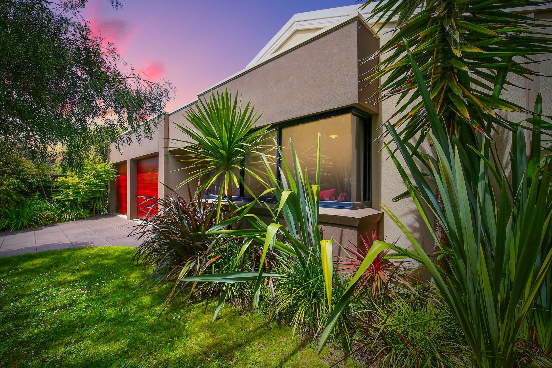 Modern house with large windows, lush tropical plants, and a green lawn at sunset