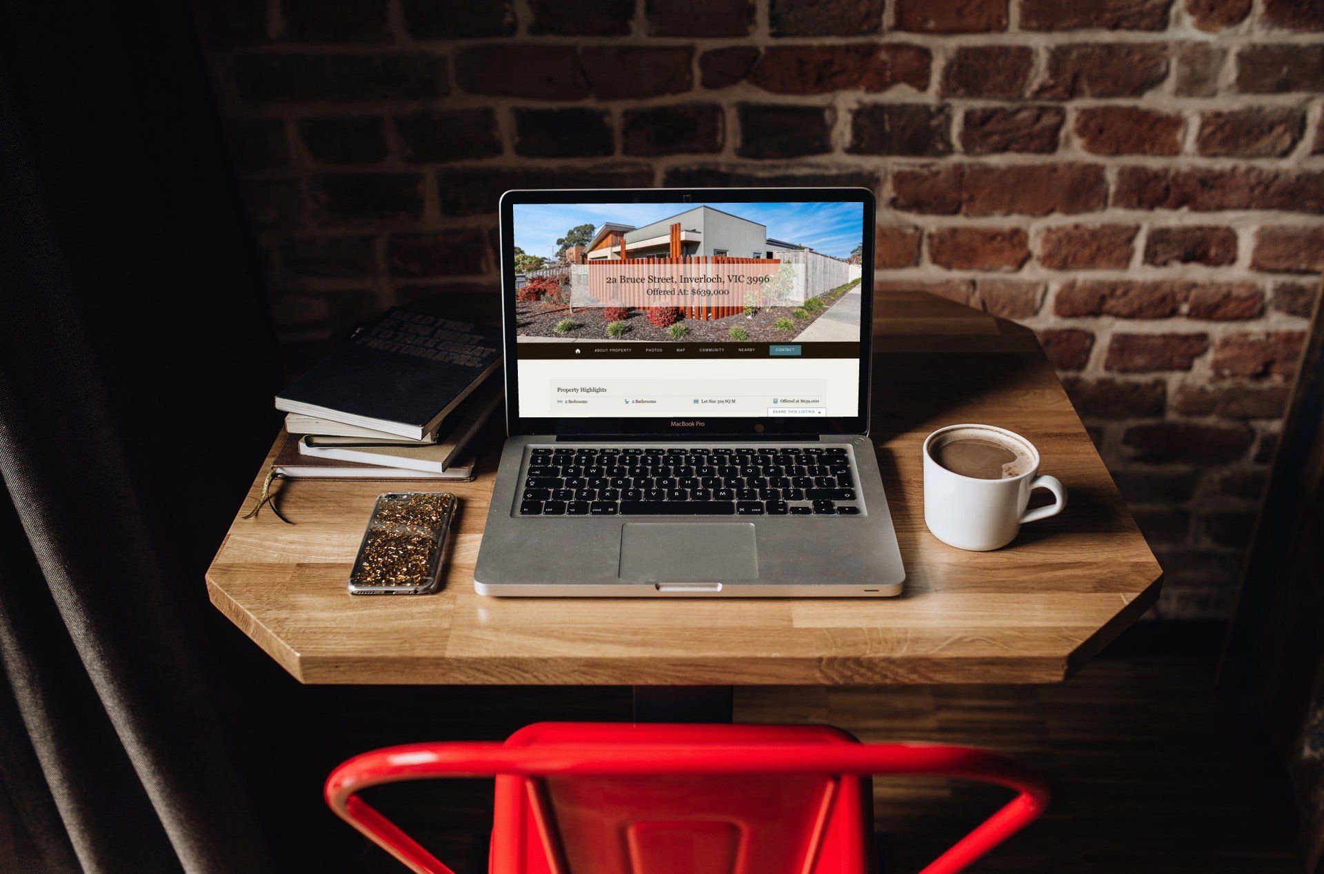 Laptop on a wooden desk in a brick-walled nook, with coffee mug, notebook, and red chair.