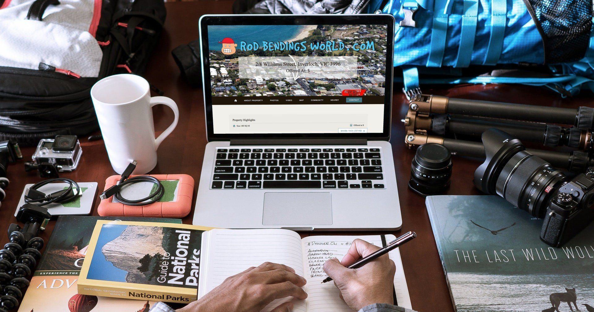 Desk with laptop, camera gear, notebook, coffee mug, and hands writing notes.