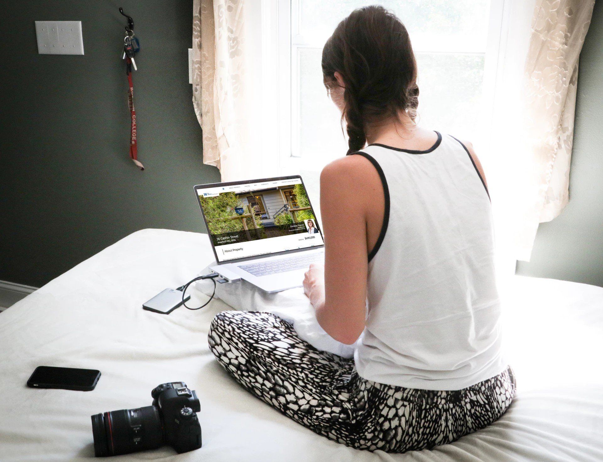 Person sitting on a bed using a laptop, with a camera and phone nearby by a bright window.