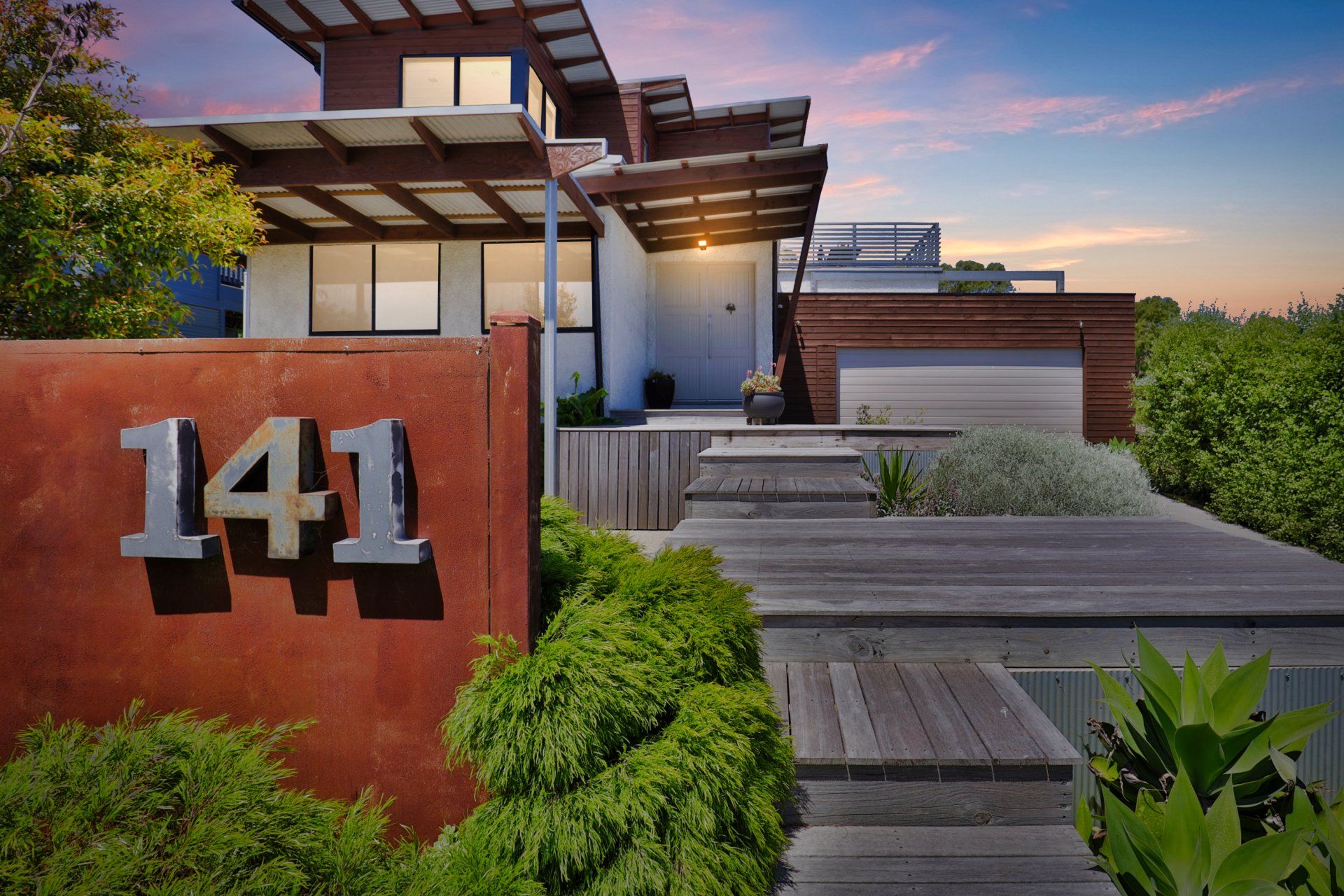 Modern house entrance with large “141” on a rust-colored wall and a wooden walkway amid greenery