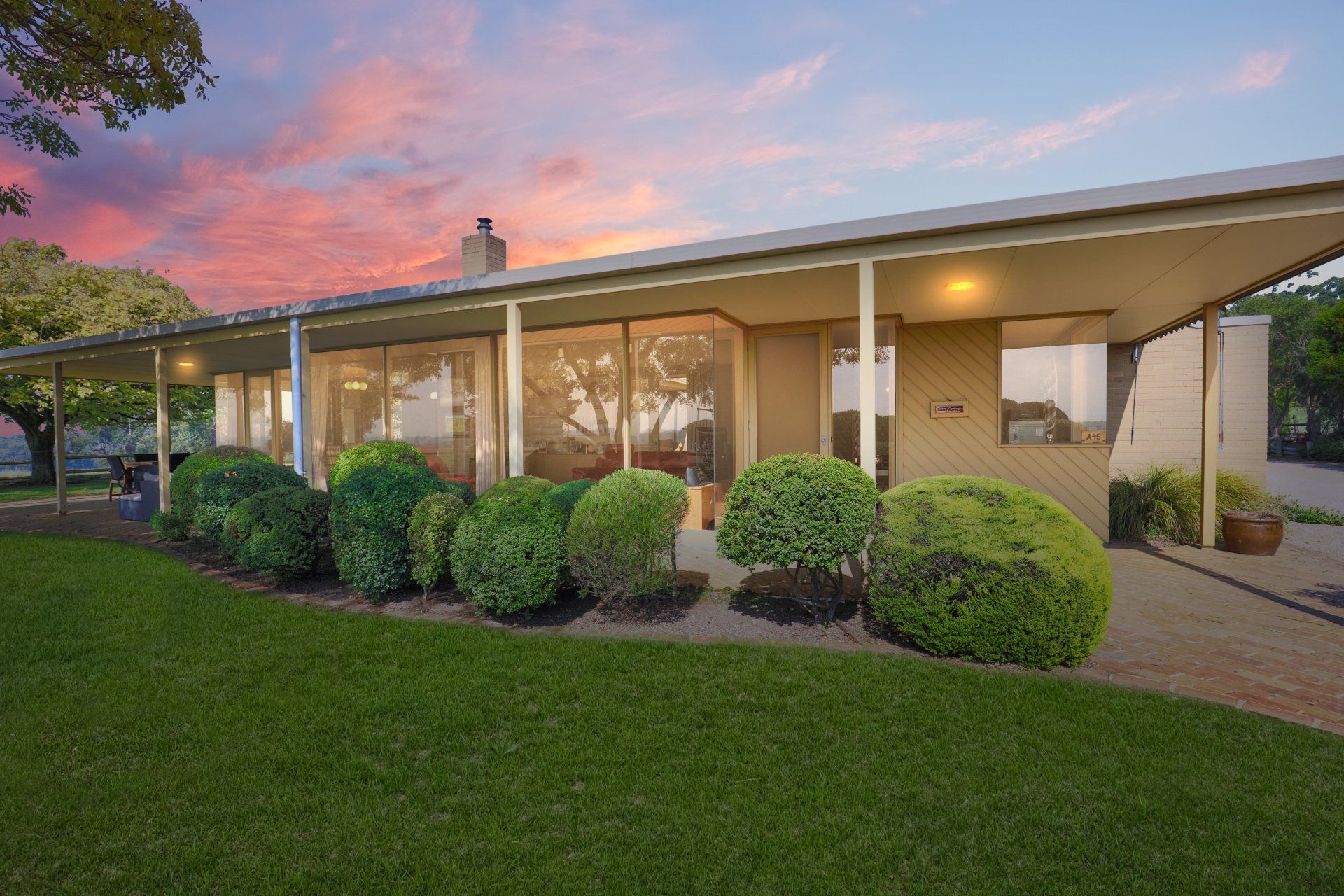 Single-story house with warm lights, manicured shrubs, and a pink sunset sky