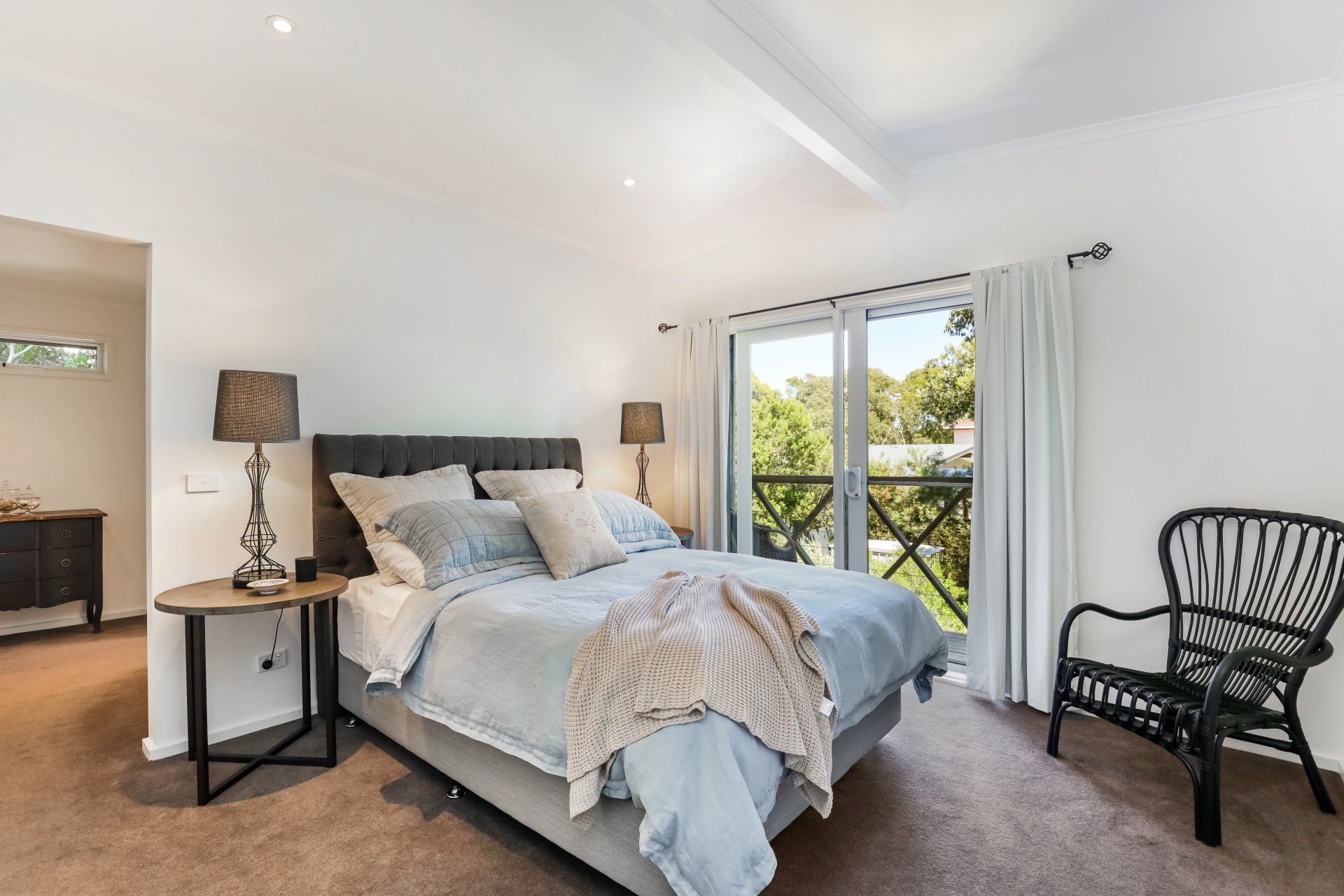 Bright bedroom with queen bed, bedside lamps, wicker chair, and balcony doors to greenery