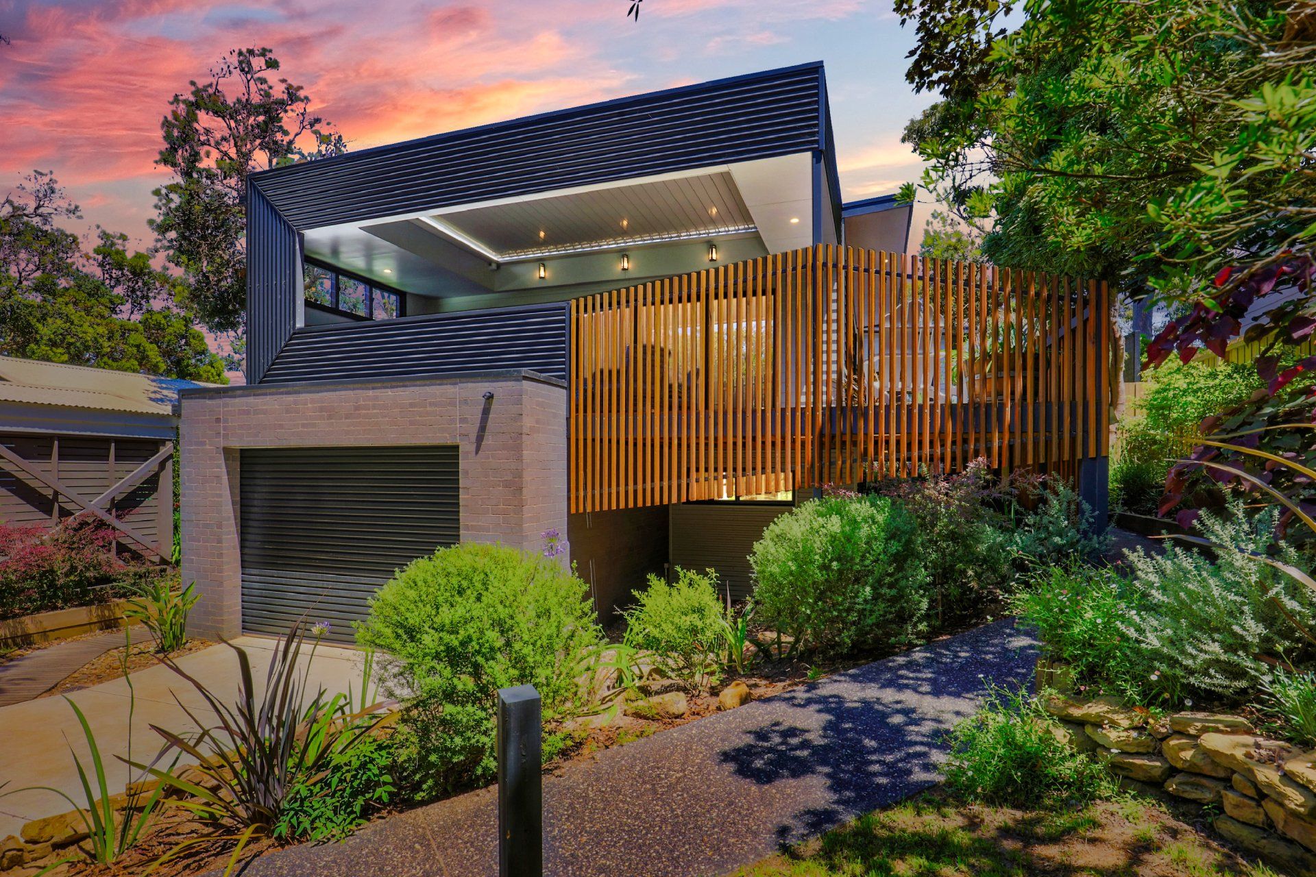 Modern house at dusk with wood slats, garage, and lush landscaping under a pink sky