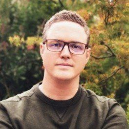 Man wearing glasses, olive green sweater, standing outside with green and yellow foliage background.