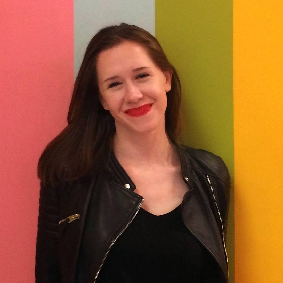 Woman in black jacket and red lipstick smiles, leaning against colorful wall.