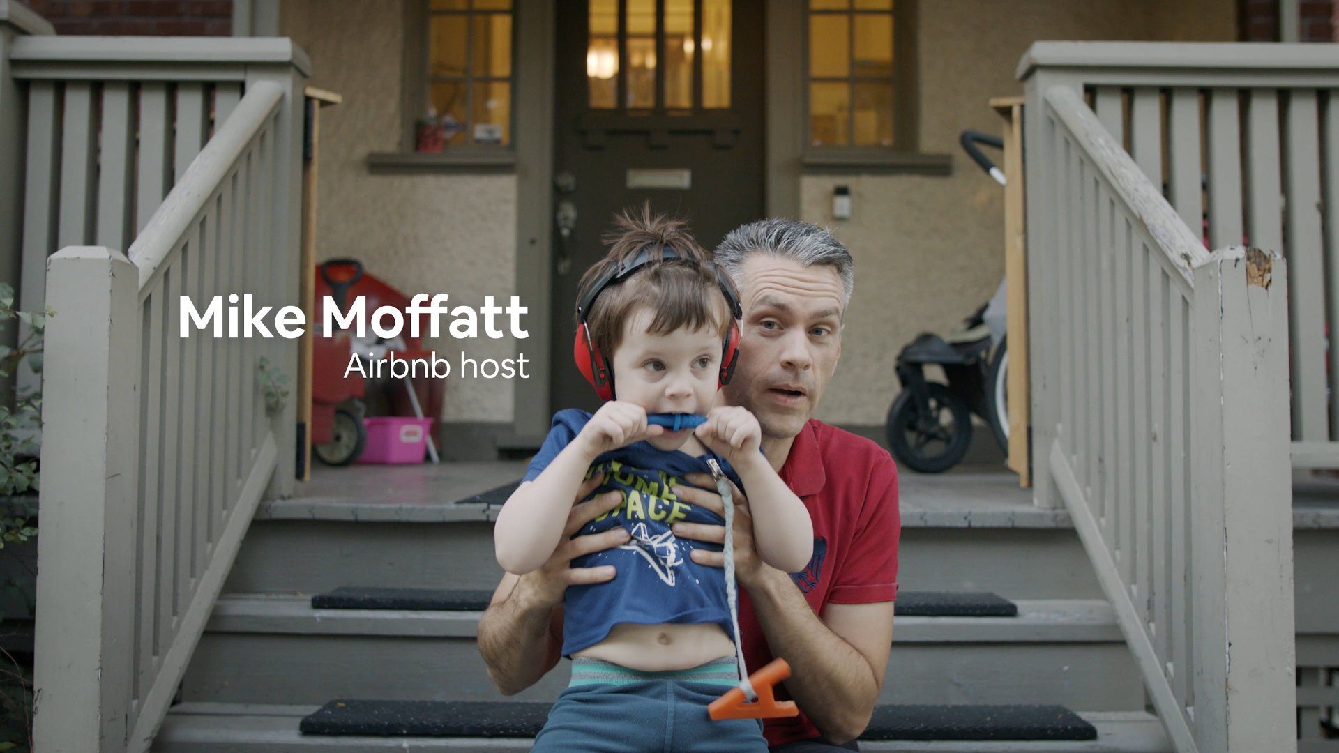 Man holding a child on steps. Man wears red shirt and looks concerned. Child has a pacifier and headphones.