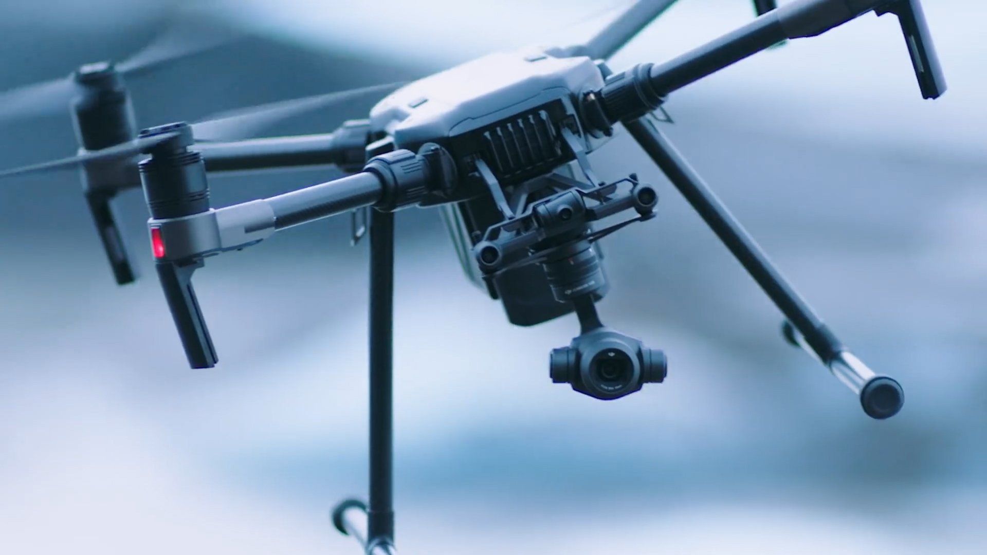 Drone in mid-air, black body, camera gimbal, propellers, landing gear, against blurred background.