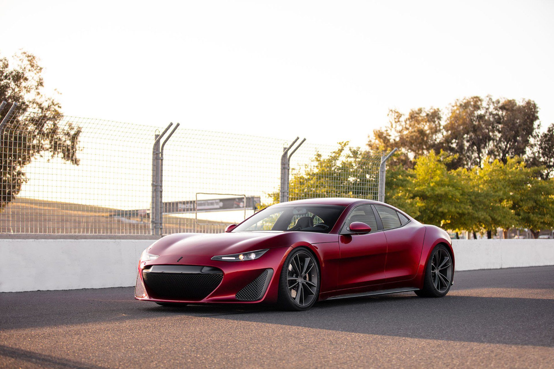 Red sports car on a racetrack, with a fence and trees in the background.