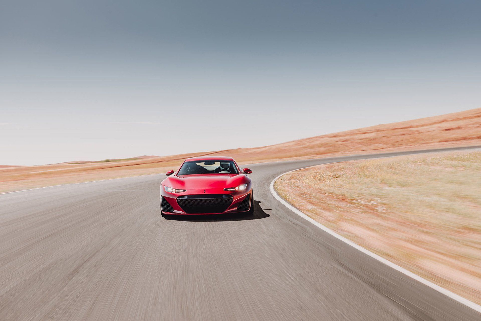 Red sports car speeding on a racetrack; sunny day, brown hills in background.