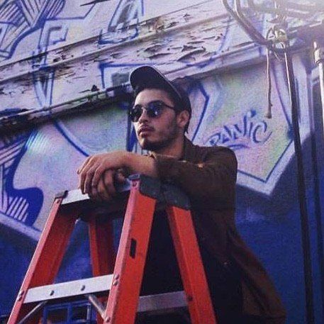 Man in sunglasses and cap leaning on an orange ladder, against a graffiti-covered wall.