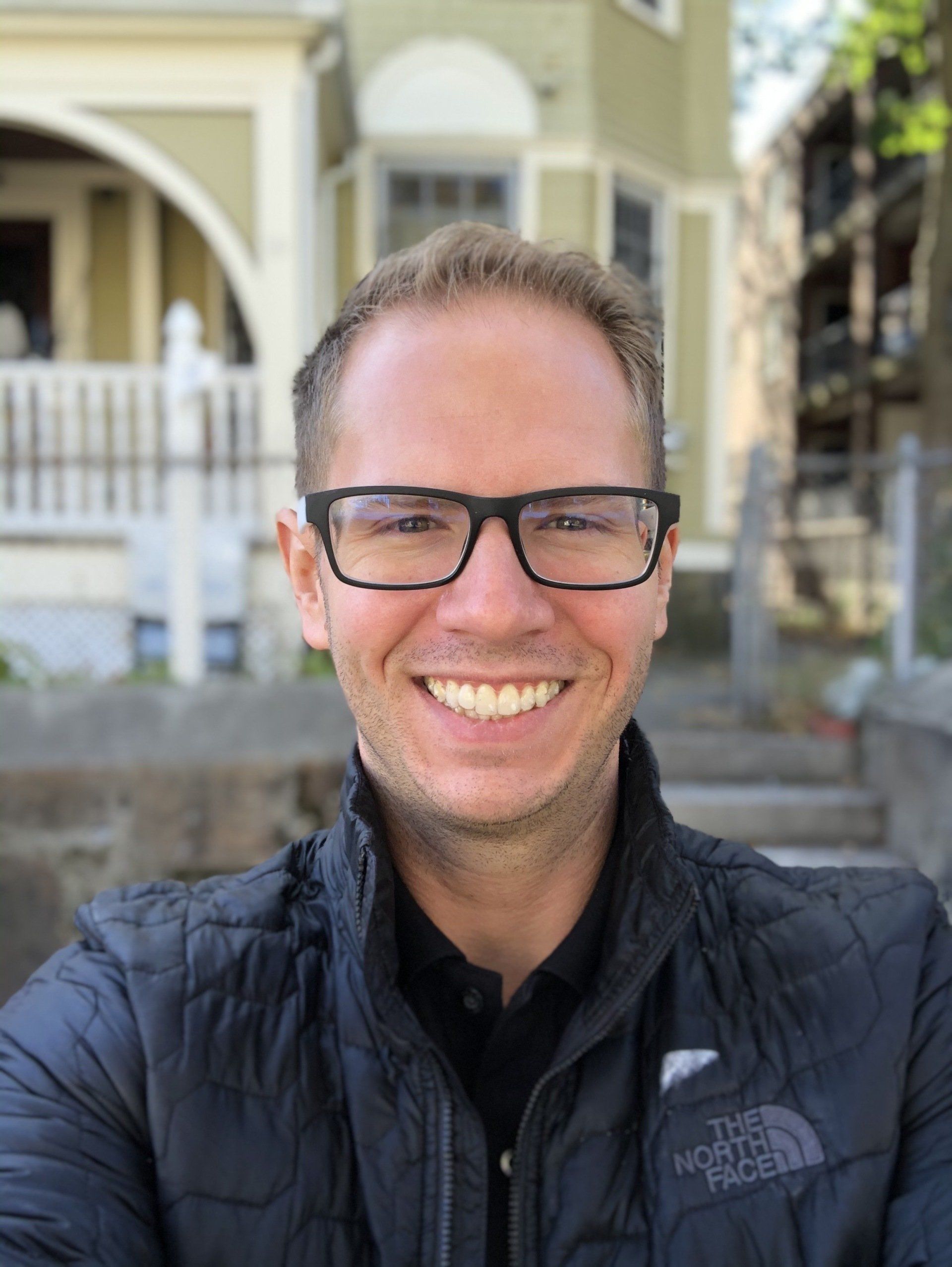 Man smiling, wearing glasses and a black jacket, standing outdoors in front of a building.