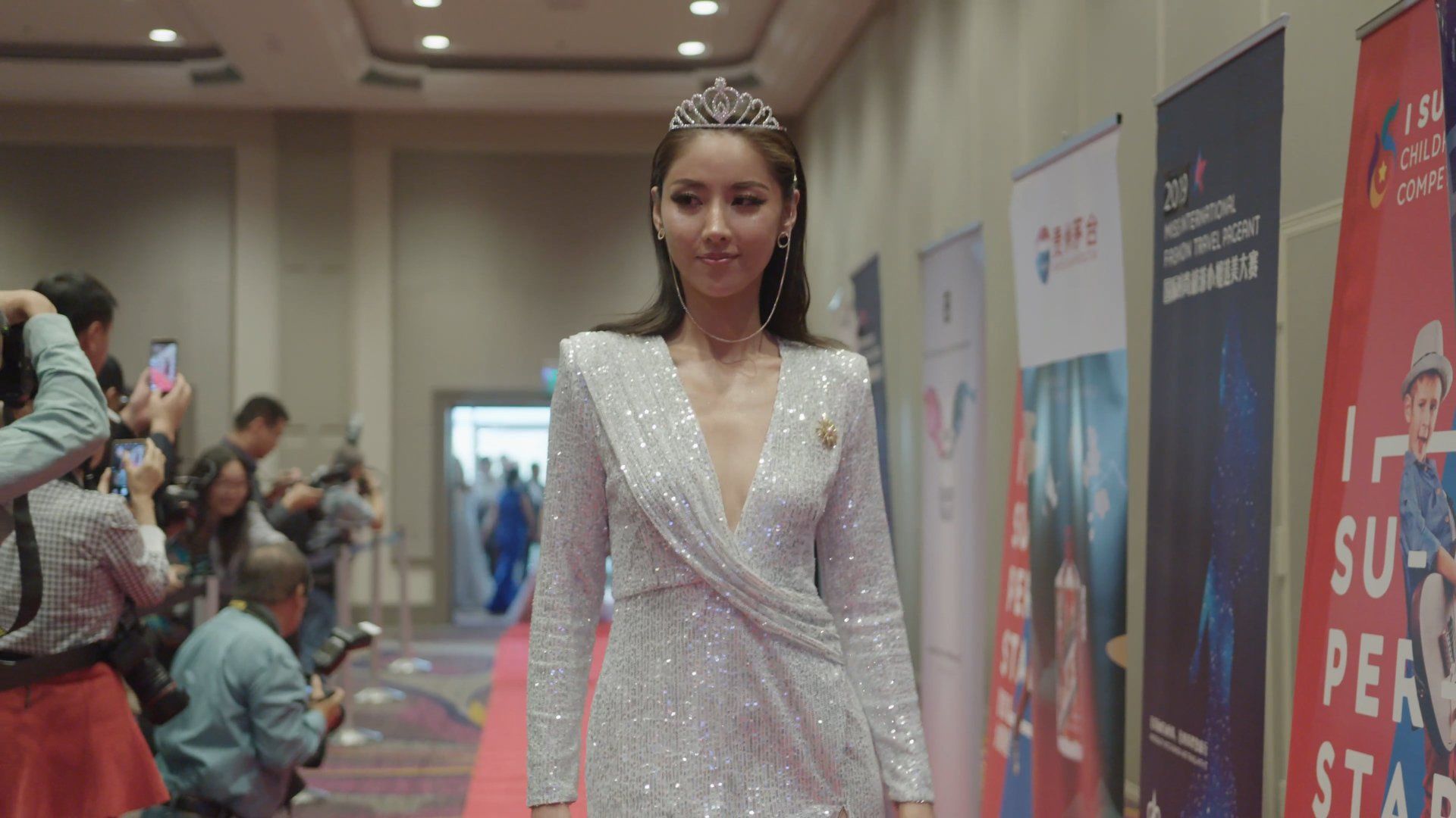 Woman in a sparkly gown and tiara walks a red carpet, smiling, flanked by people taking photos.