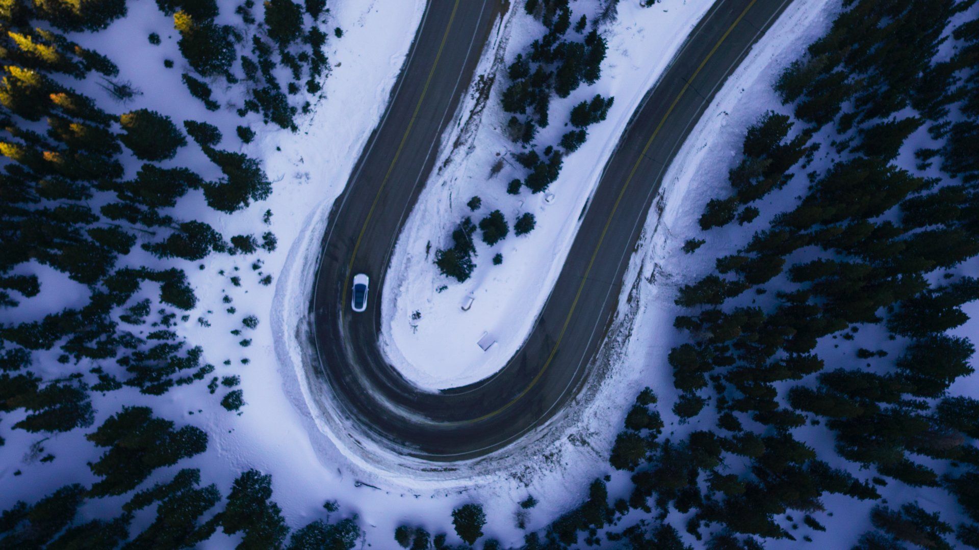 Aerial view of a car on a winding road through a snowy mountain pass, surrounded by evergreen trees.