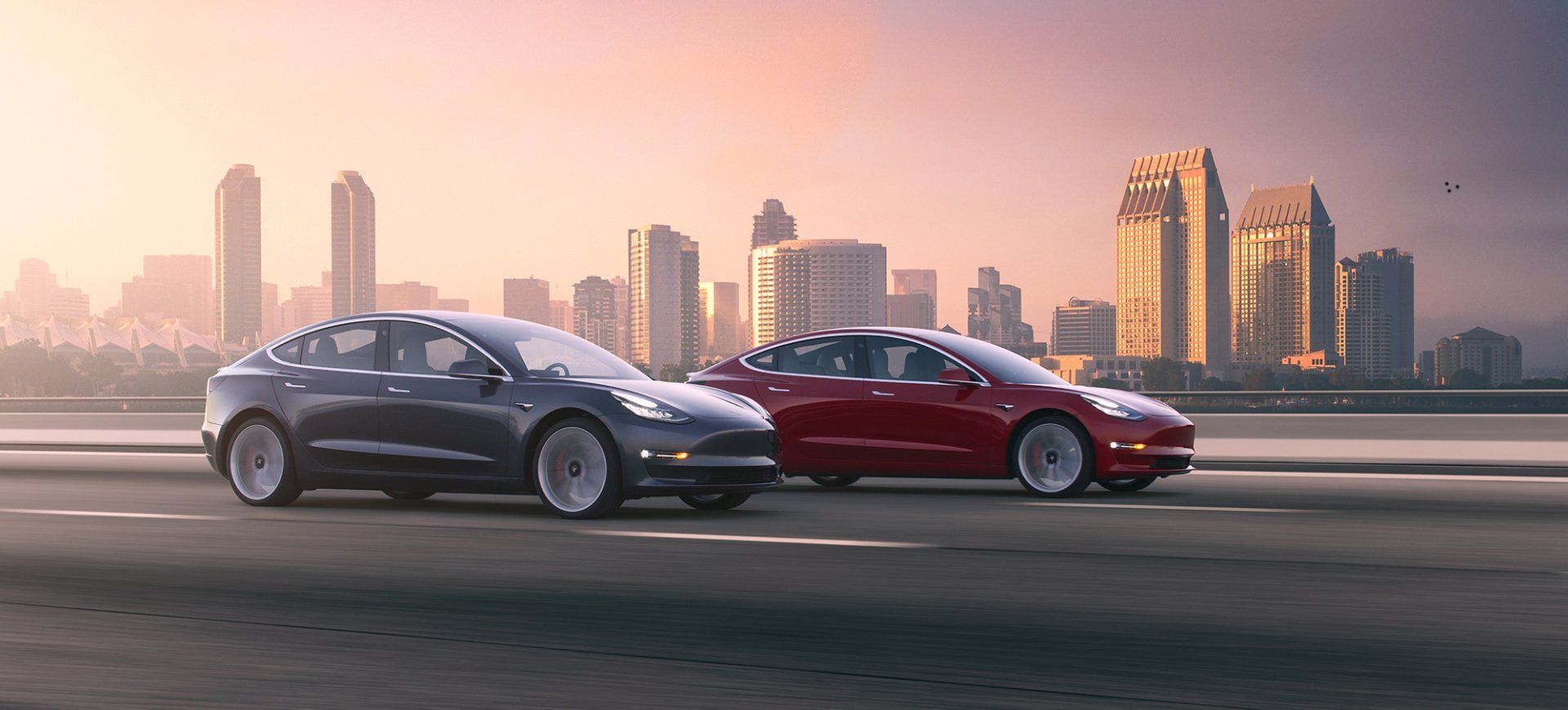 Two Tesla Model 3 cars racing on a road with a city skyline in the background, lit by a sunset.