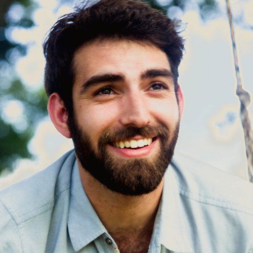 Man with dark hair and beard, smiling and looking upward in a light blue shirt, outdoors.