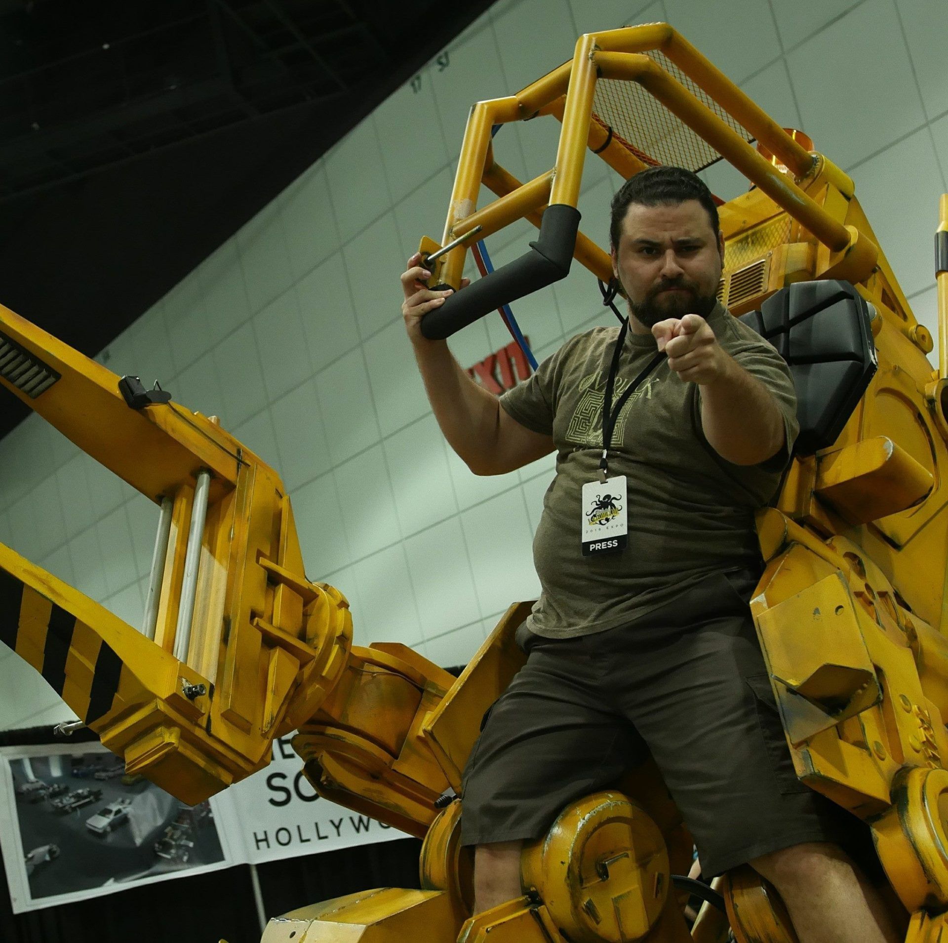 Man in khaki shirt points at viewer while seated on a yellow, mechanical exoskeleton.