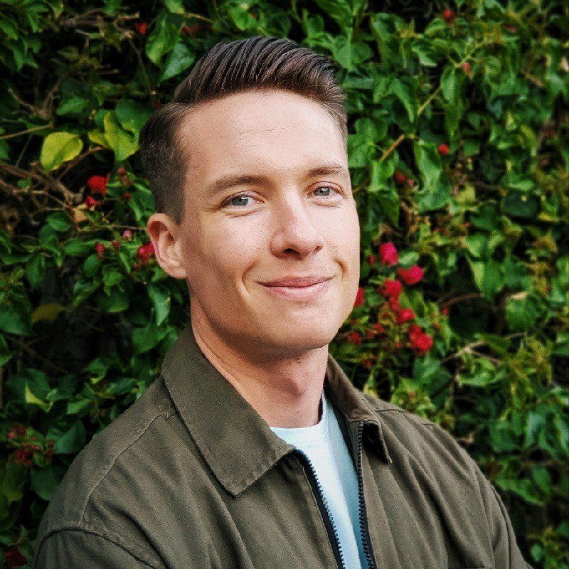 Man with short brown hair and a friendly smile, wearing a green jacket, in front of greenery.
