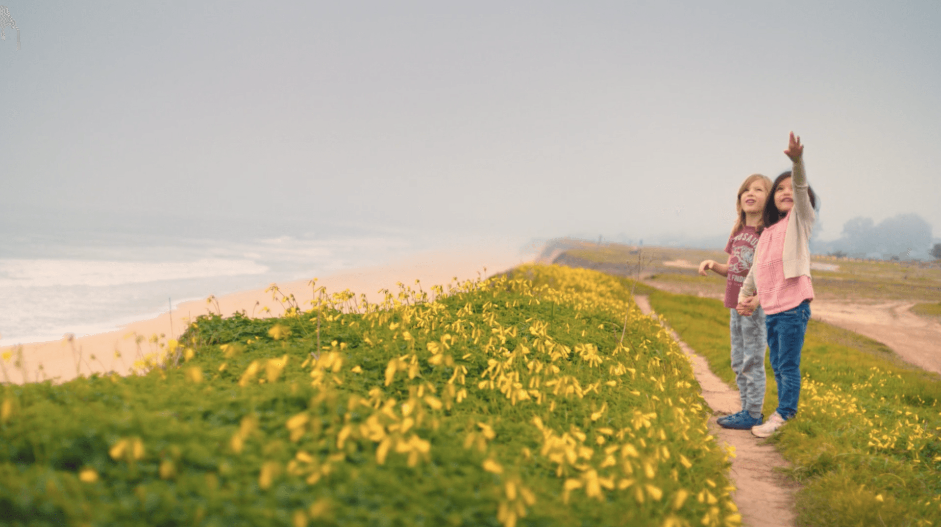 Two girls on a path near the ocean, one pointing up. Yellow flowers line the path.